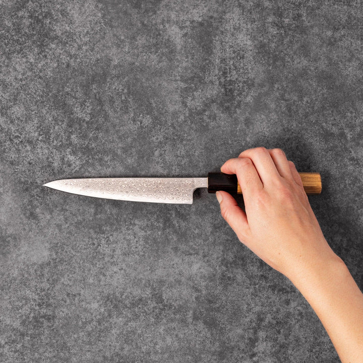 Japanese Damascus petty knife with intricate layered steel blade and natural wood handle placed on gray stone surface.