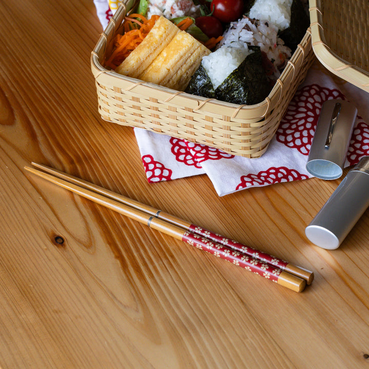 Sakura portable Japanese travel chopsticks with red flower design next to a bamboo bento box filled with onigiri