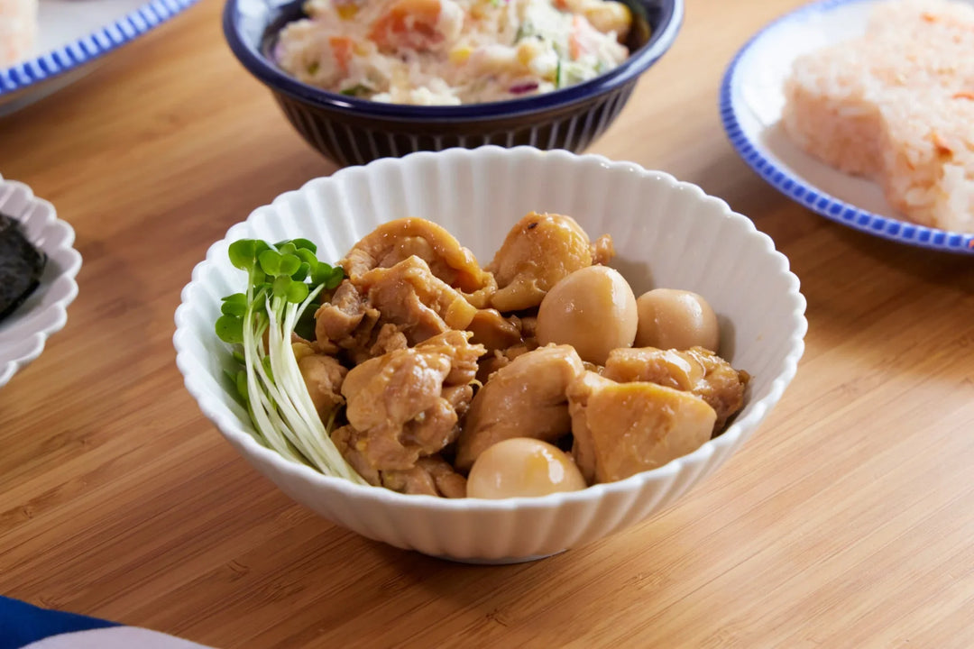 A white scalloped bowl filled with simmered chicken and quail eggs, served with microgreens on a wooden table during a home meal.