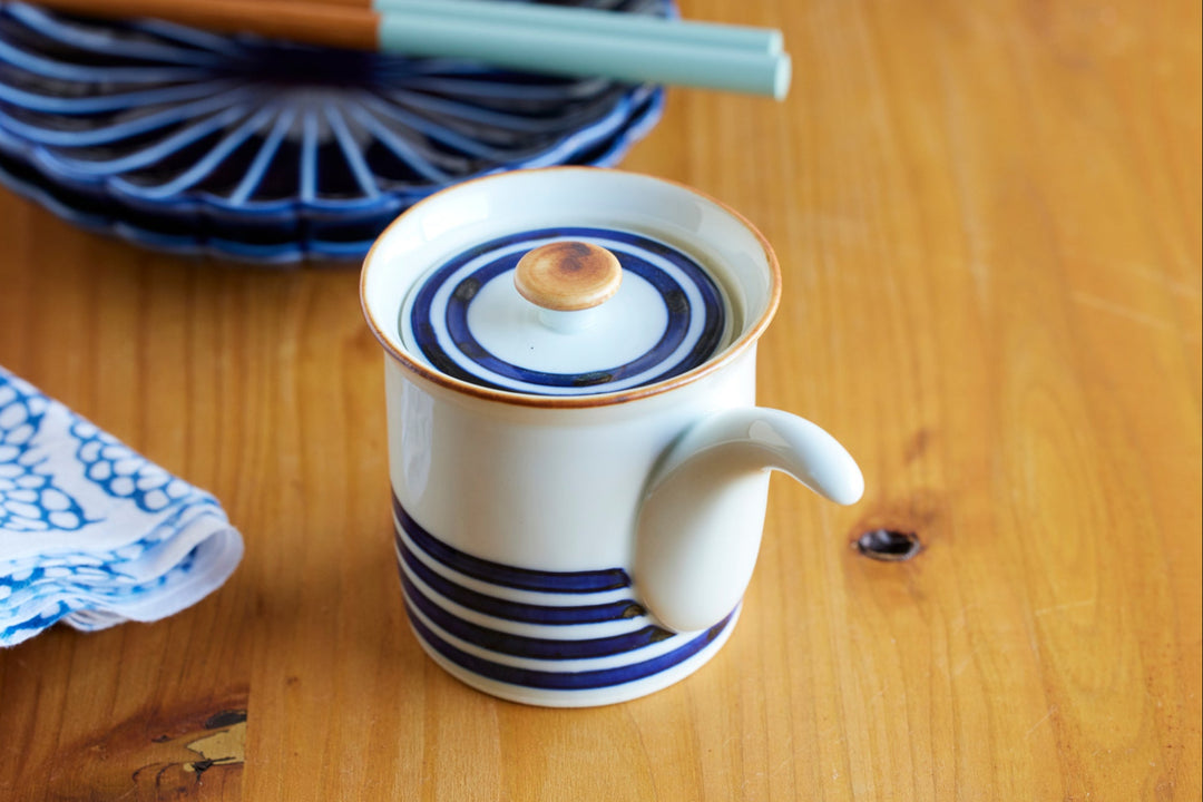 White ceramic soy sauce dispenser with a blue stripe design and curved spout placed on a wooden table beside blue tableware.