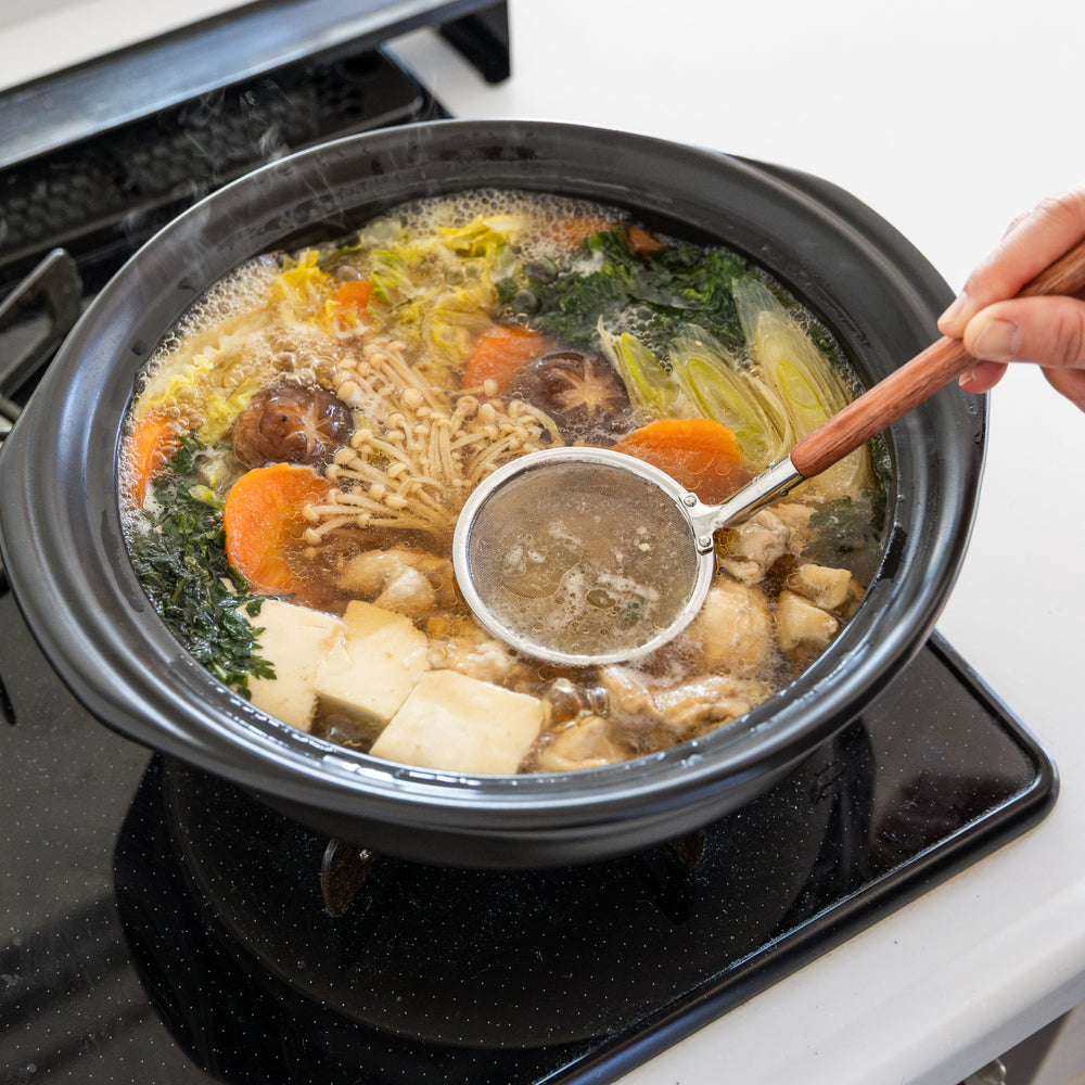 Stainless steel kitchen skimmer with wooden handle removing foam from simmering donabe hot pot