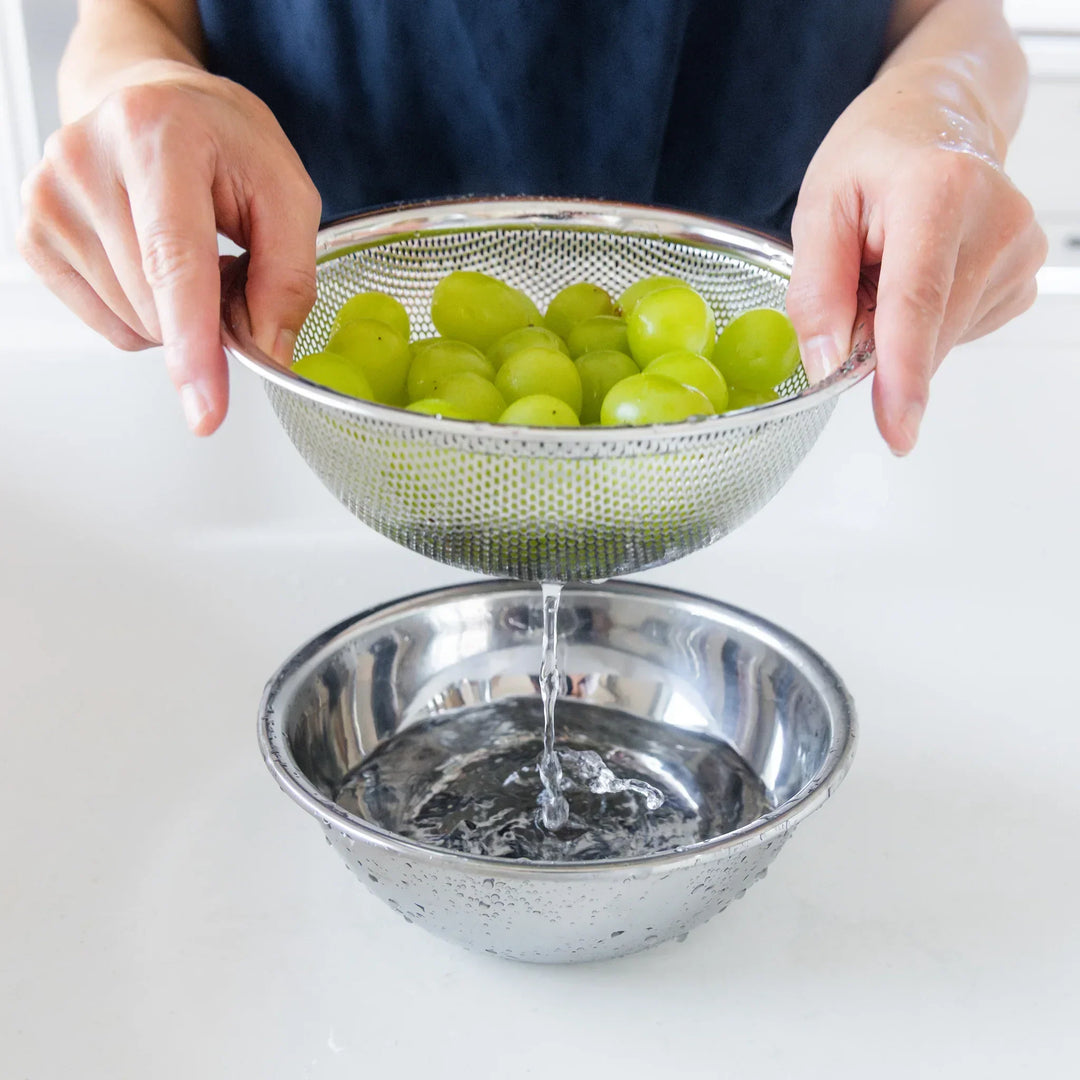 Nami rinsing green grapes in a metal colander over a silver bowl on a white surface.