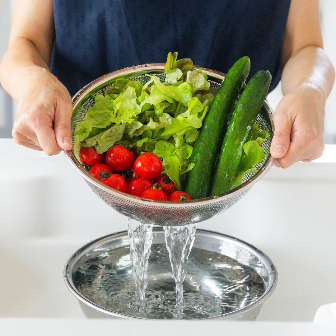 Freshly washed vegetables in a stainless steel mesh strainer set over a matching bowl, draining water after rinsing veggies.
