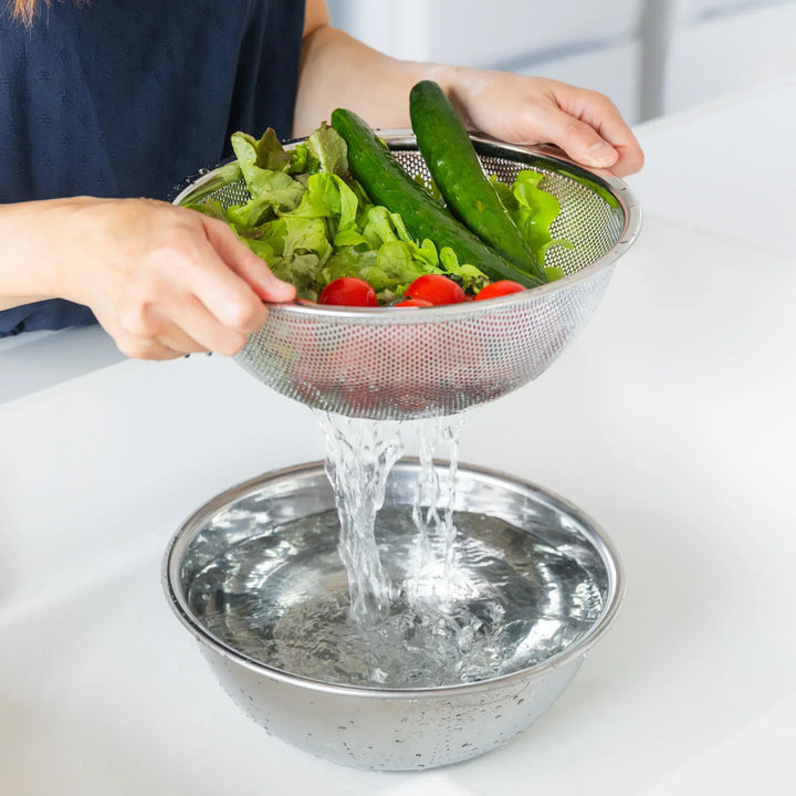 Nami washing vegetables in a metal colander over a larger bowl on a white surface.