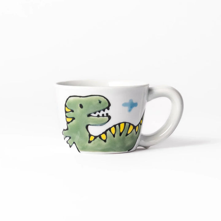 White ceramic kids mug featuring green T-Rex dinosaur graphic with yellow accents and blue cloud motifs, round handle