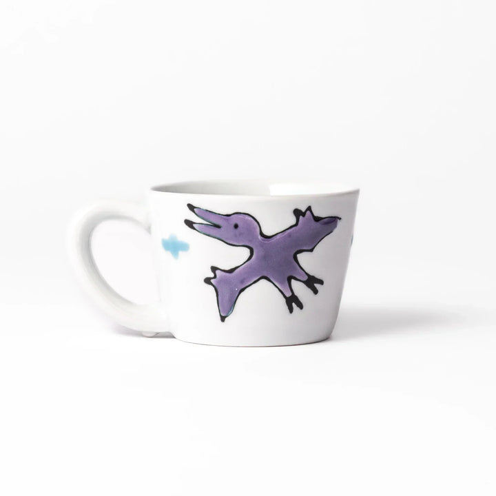 White ceramic kids mug featuring green T-Rex dinosaur graphic with yellow accents and blue cloud motifs, round handle