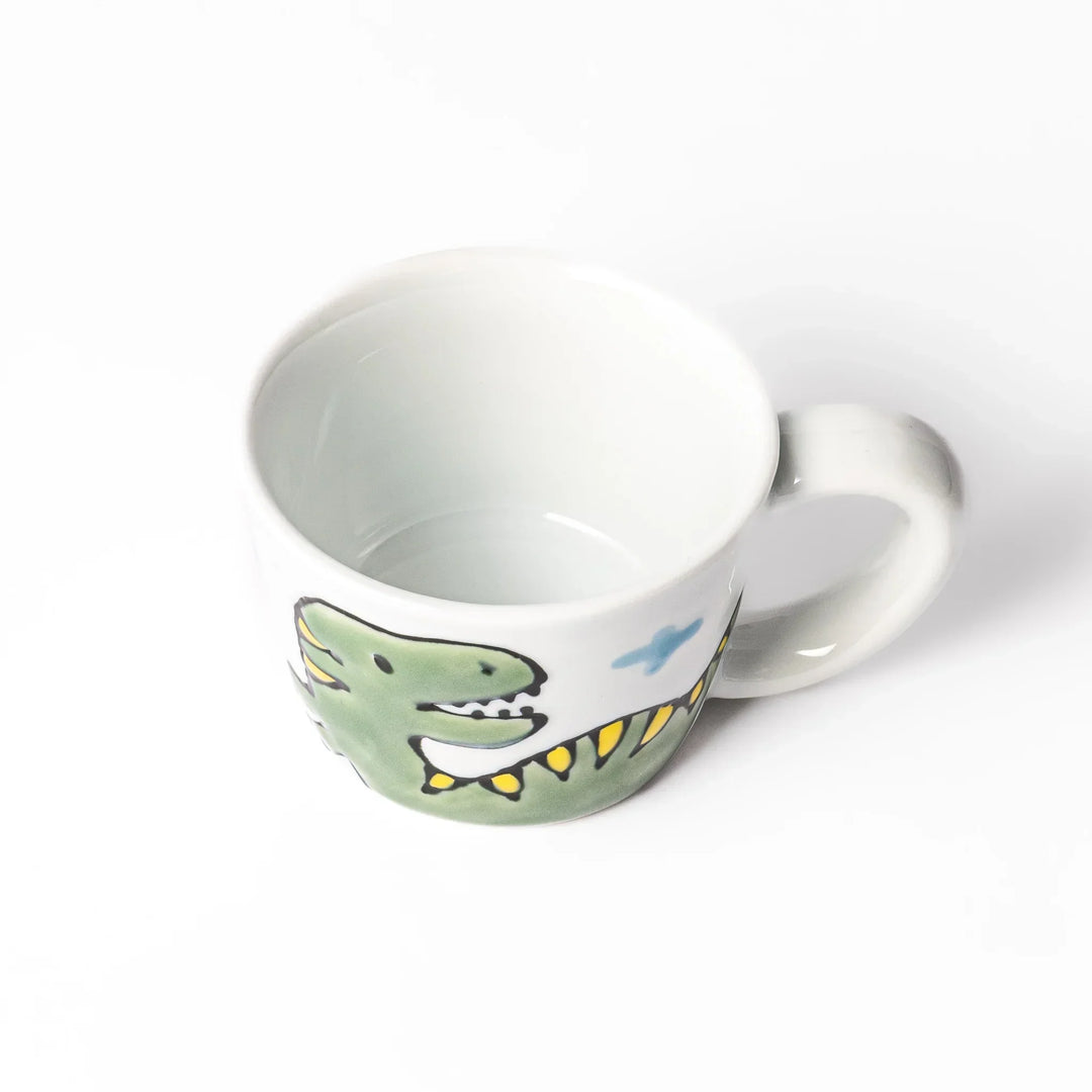 White ceramic kids mug featuring green T-Rex dinosaur graphic with yellow accents and blue cloud motifs, round handle