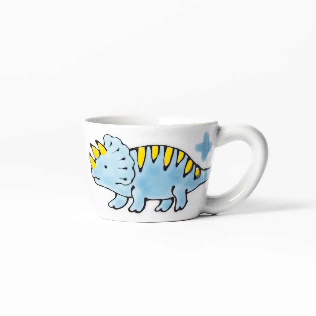 White ceramic kids mug with blue triceratops graphic, yellow horns and stripes, plus blue cloud accents, round handle.