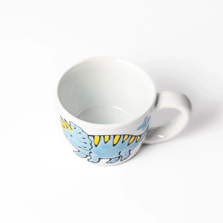 White ceramic kids mug with blue triceratops graphic, yellow horns and stripes, plus blue cloud accents, round handle.