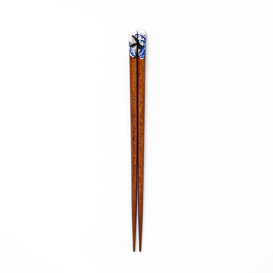 Pair of brown wooden chopsticks with white ceramic tips featuring a blue wave and whale design on a white background.
