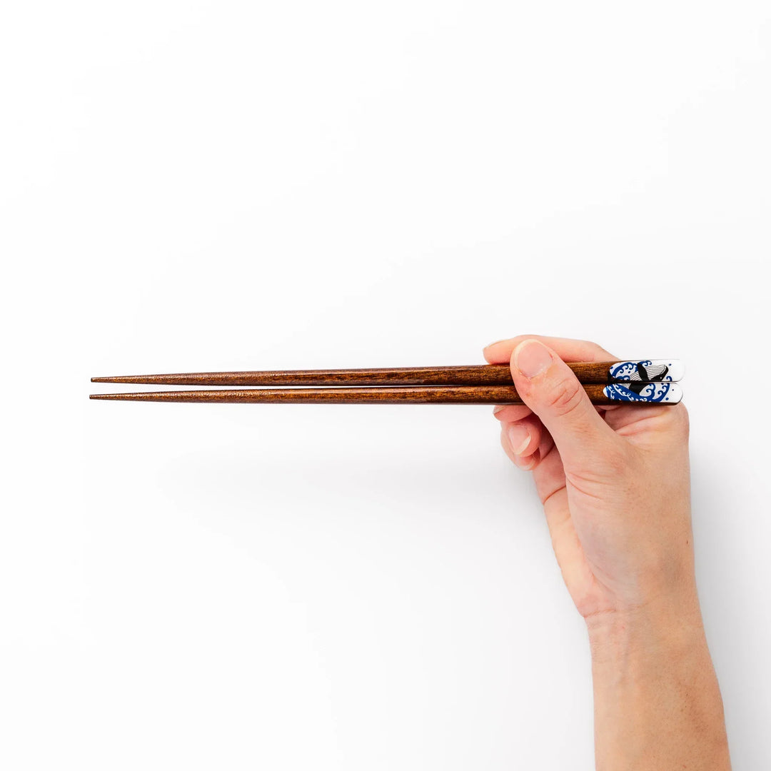 Pair of brown wooden chopsticks with white ceramic tips featuring a blue wave and whale design on a white background.