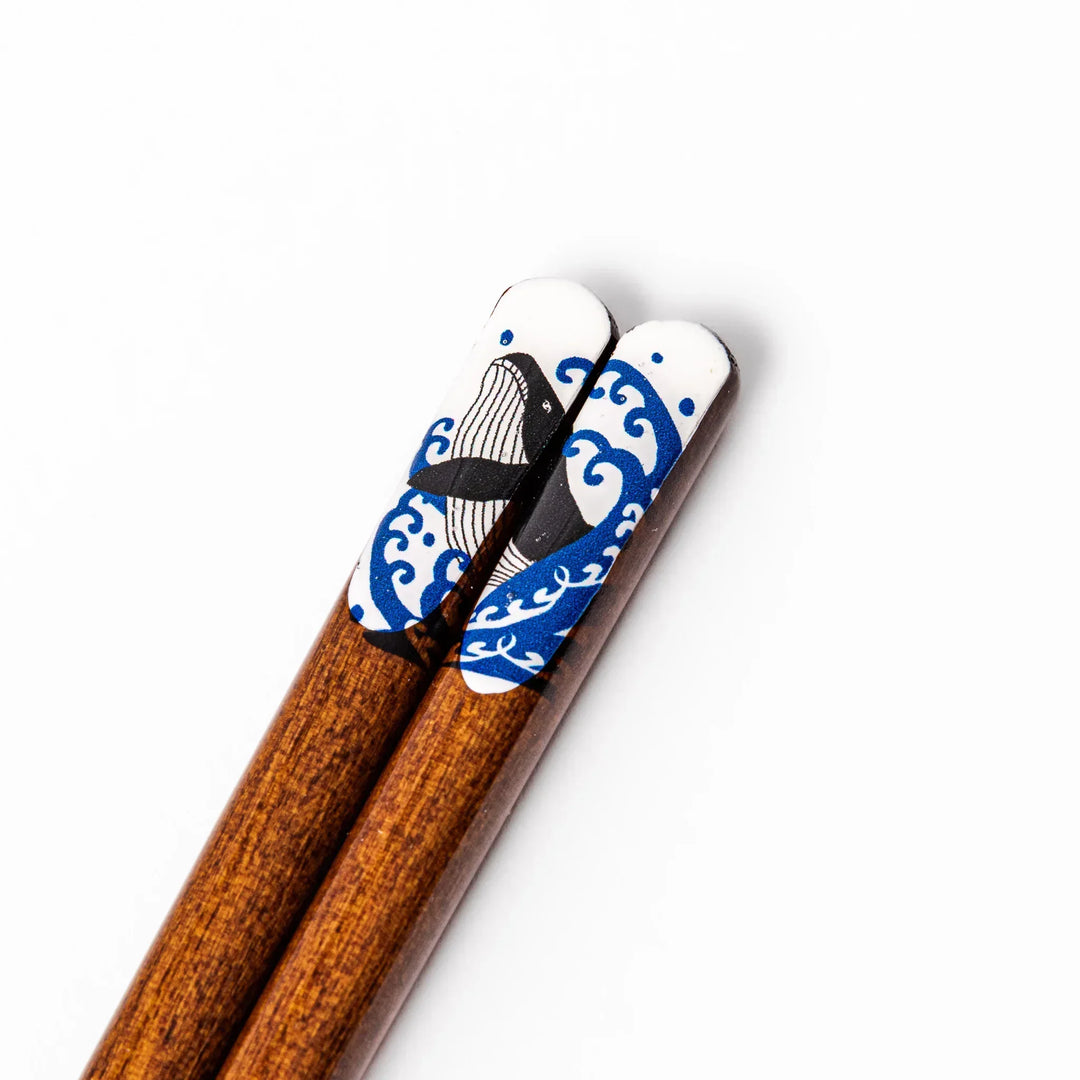 Pair of brown wooden chopsticks with white ceramic tips featuring a blue wave and whale design on a white background.
