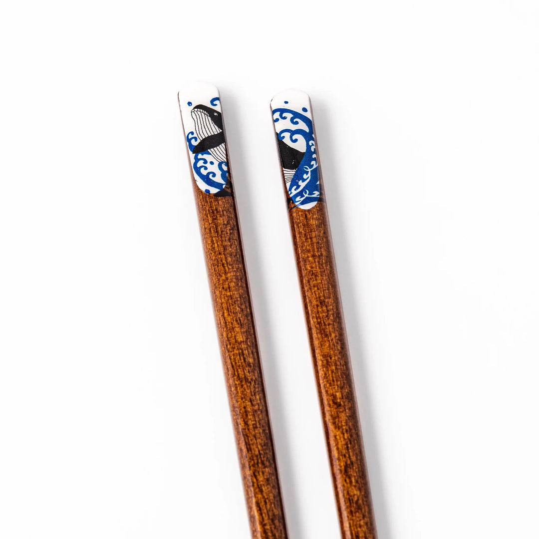 Pair of brown wooden chopsticks with white ceramic tips featuring a blue wave and whale design on a white background.