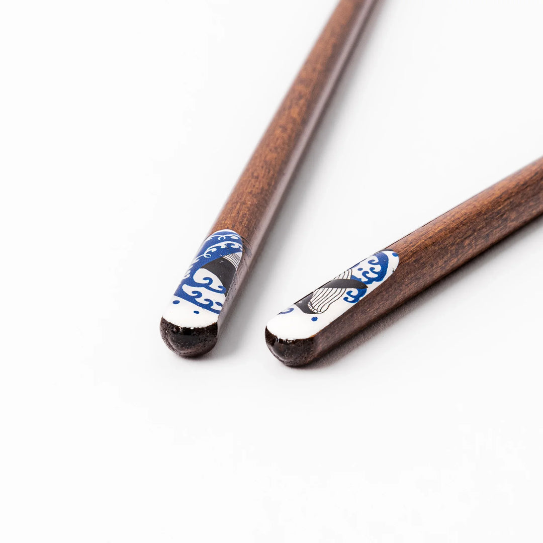 Pair of brown wooden chopsticks with white ceramic tips featuring a blue wave and whale design on a white background.