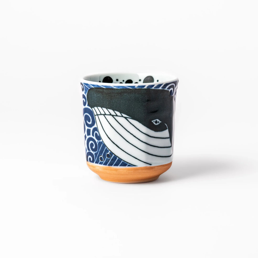 Porcelain tea cup featuring a bold black-and-white whale design surrounded by blue ocean waves with an unglazed brown base.