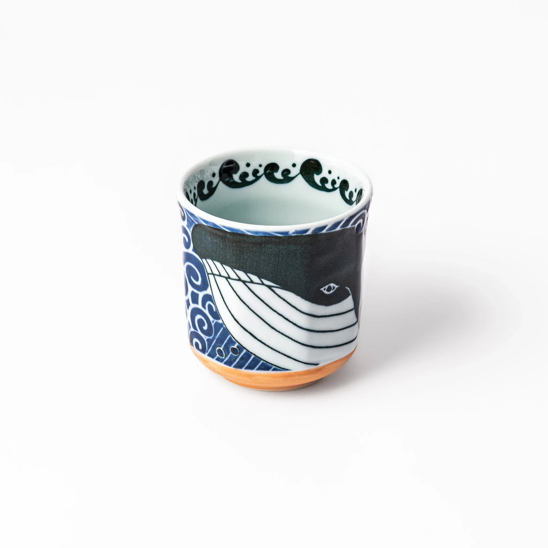 Porcelain tea cup featuring a bold black-and-white whale design surrounded by blue ocean waves with an unglazed brown base.
