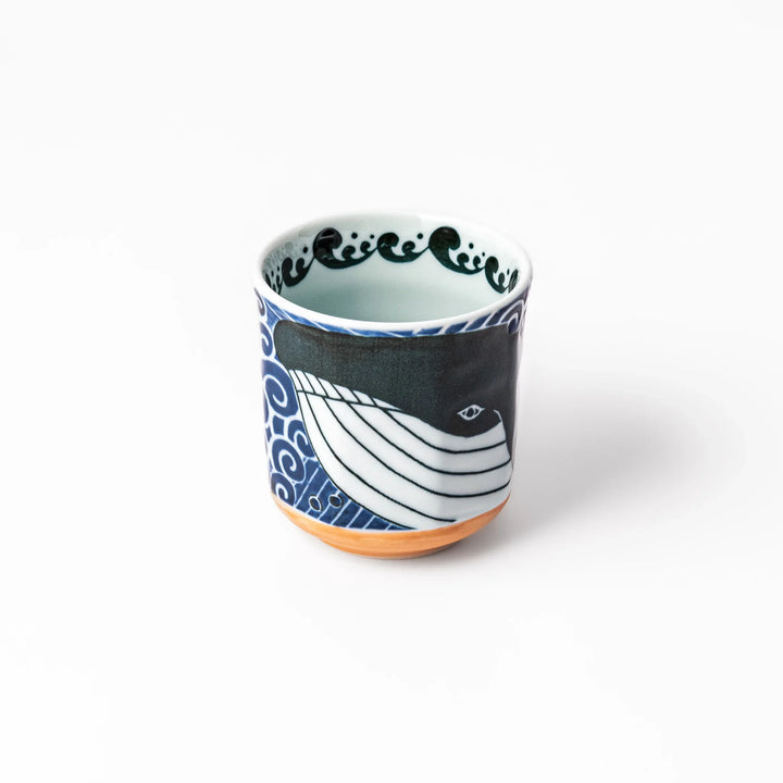 Porcelain tea cup featuring a bold black-and-white whale design surrounded by blue ocean waves with an unglazed brown base.