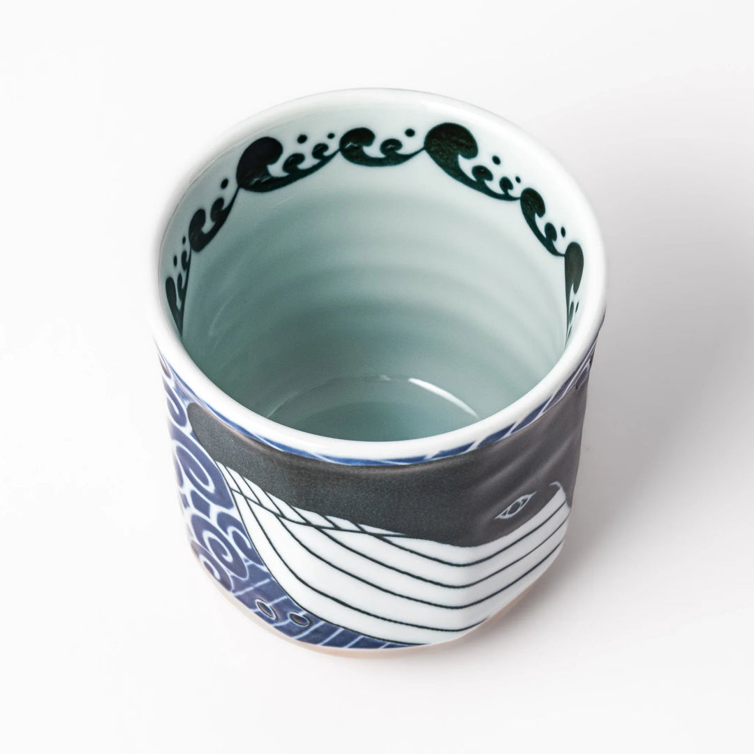 Porcelain tea cup featuring a bold black-and-white whale design surrounded by blue ocean waves with an unglazed brown base.