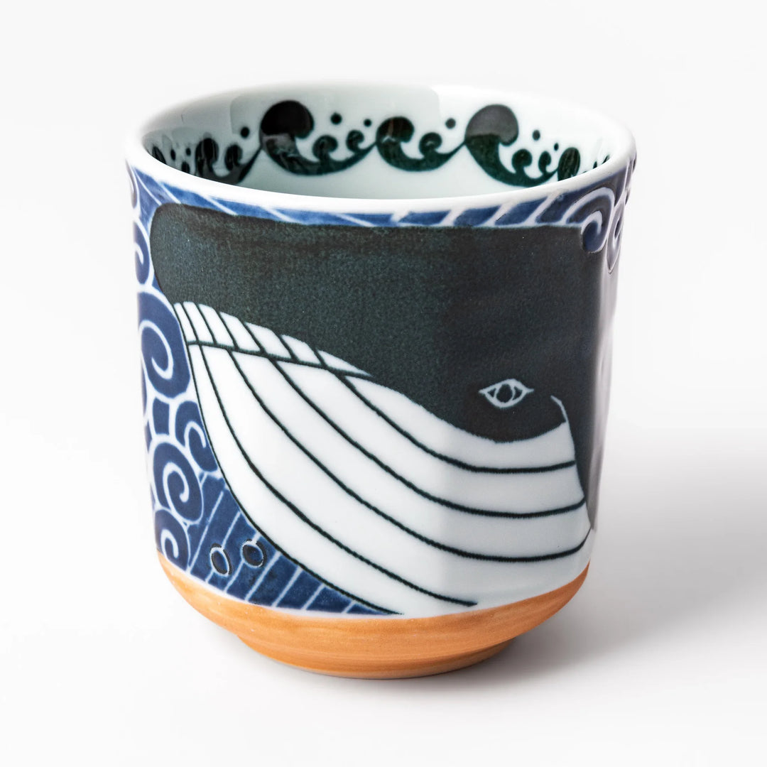 Porcelain tea cup featuring a bold black-and-white whale design surrounded by blue ocean waves with an unglazed brown base.