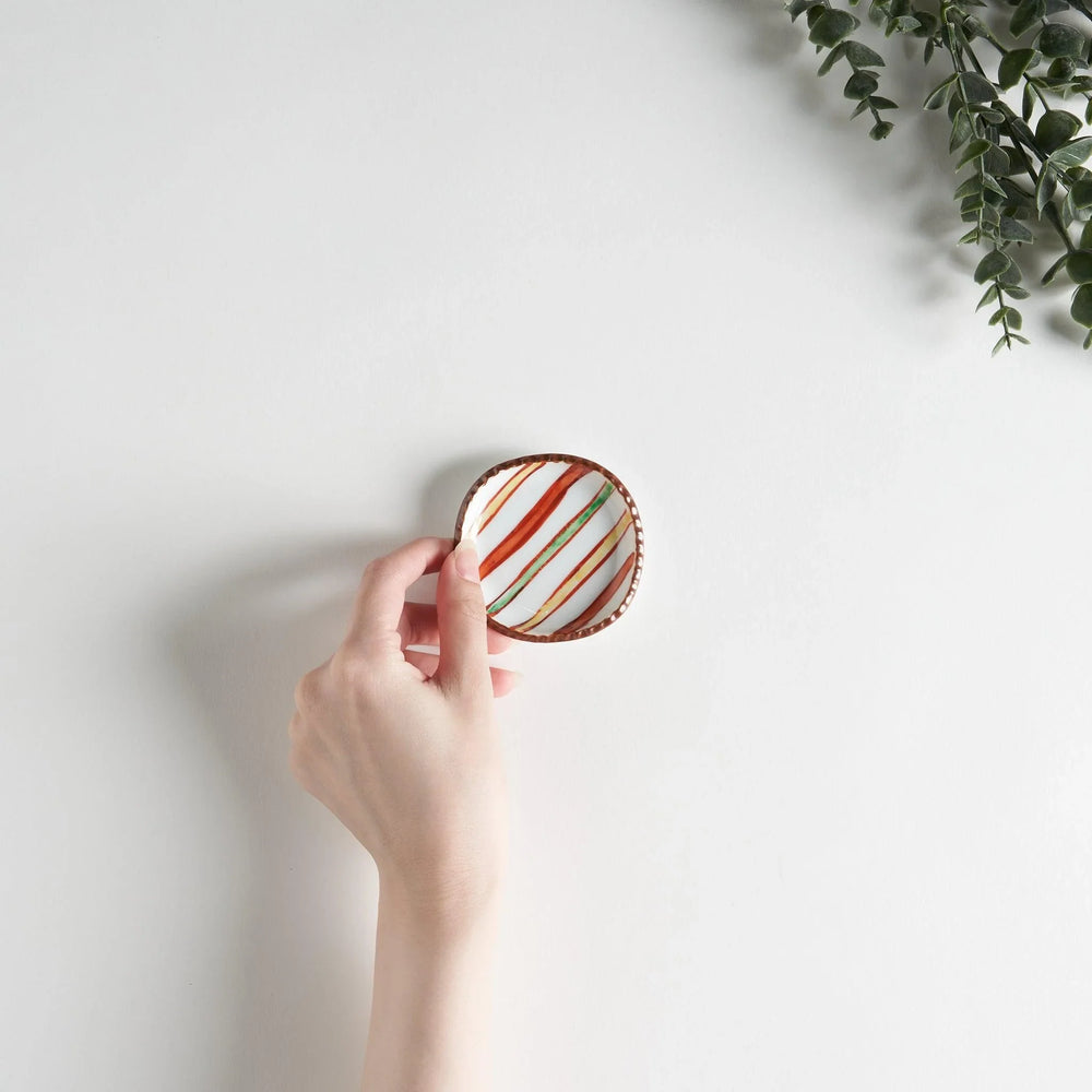 A hand holding a colorful striped sauce dish above a plain white surface, showcasing its vibrant design.