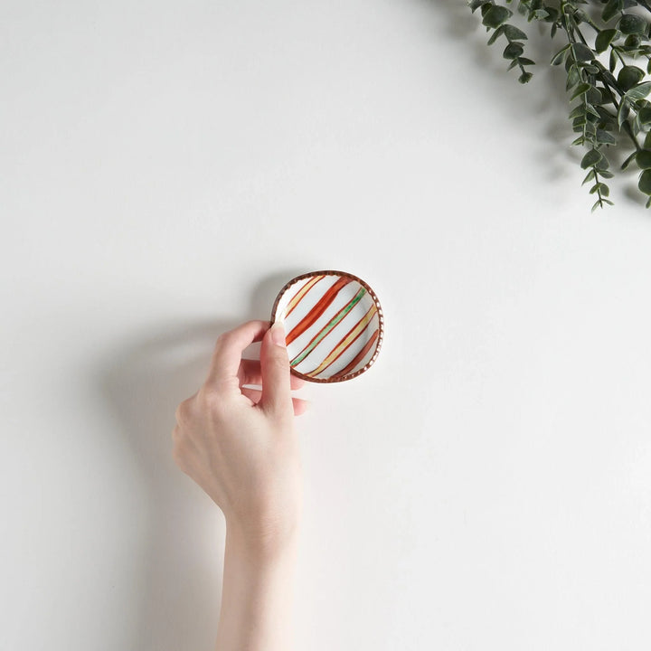 A hand holding a colorful striped sauce dish above a plain white surface, showcasing its vibrant design.