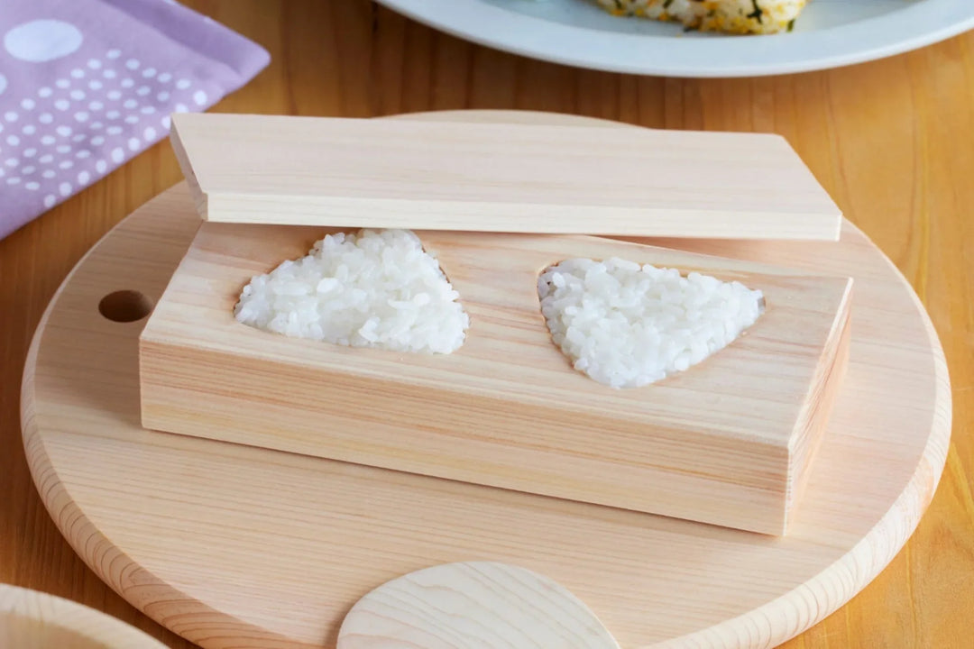 Wooden onigiri mold shaping two rice triangles on a table with rice bowl, pickles, and assorted homemade onigiri
