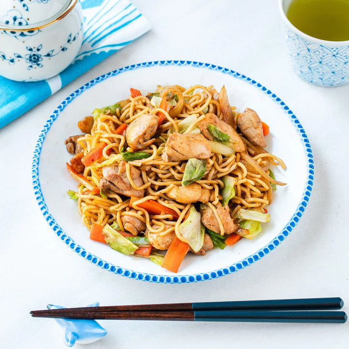 Chicken yakisoba with vegetables on white plate with blue rim served with chopsticks and green tea.