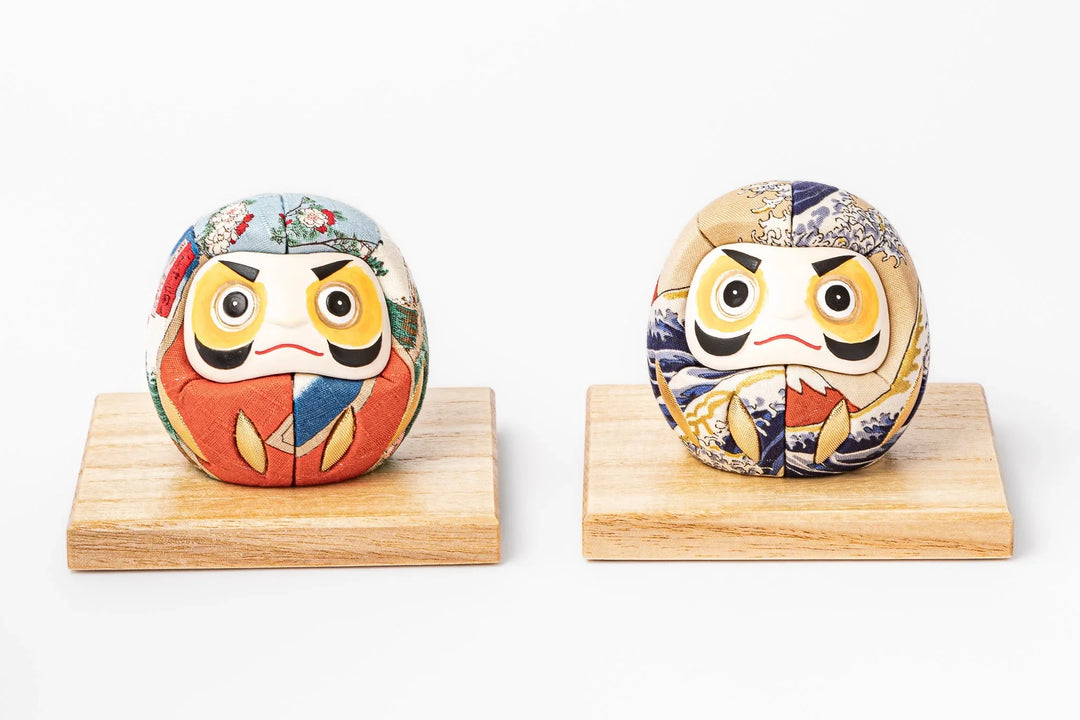 Two traditional Japanese fabric daruma dolls with colorful patterns displayed on wooden stands side by side