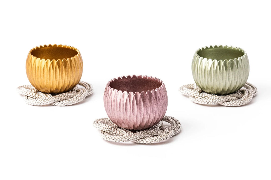 Three hydrangea shaped metal prayer bowls in gold pink and green, each set on a decorative braided coaster against a white background.