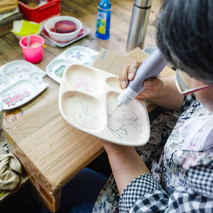 Artisan hand painting a dinosaur design onto a kids section plate during the detailed decorating process