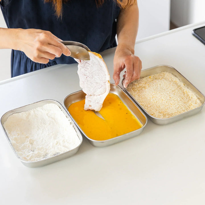 Nami dipping a flour-coated pork slice into beaten egg using stainless prep trays arranged for Japanese tonkatsu breading.