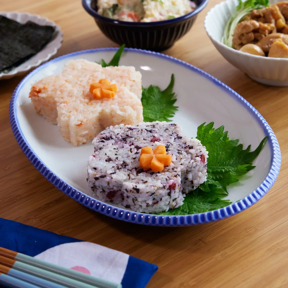 Sakura and plum blossom-shaped rice balls on a white oval plate with scalloped blue rim, garnished with shiso leaves.