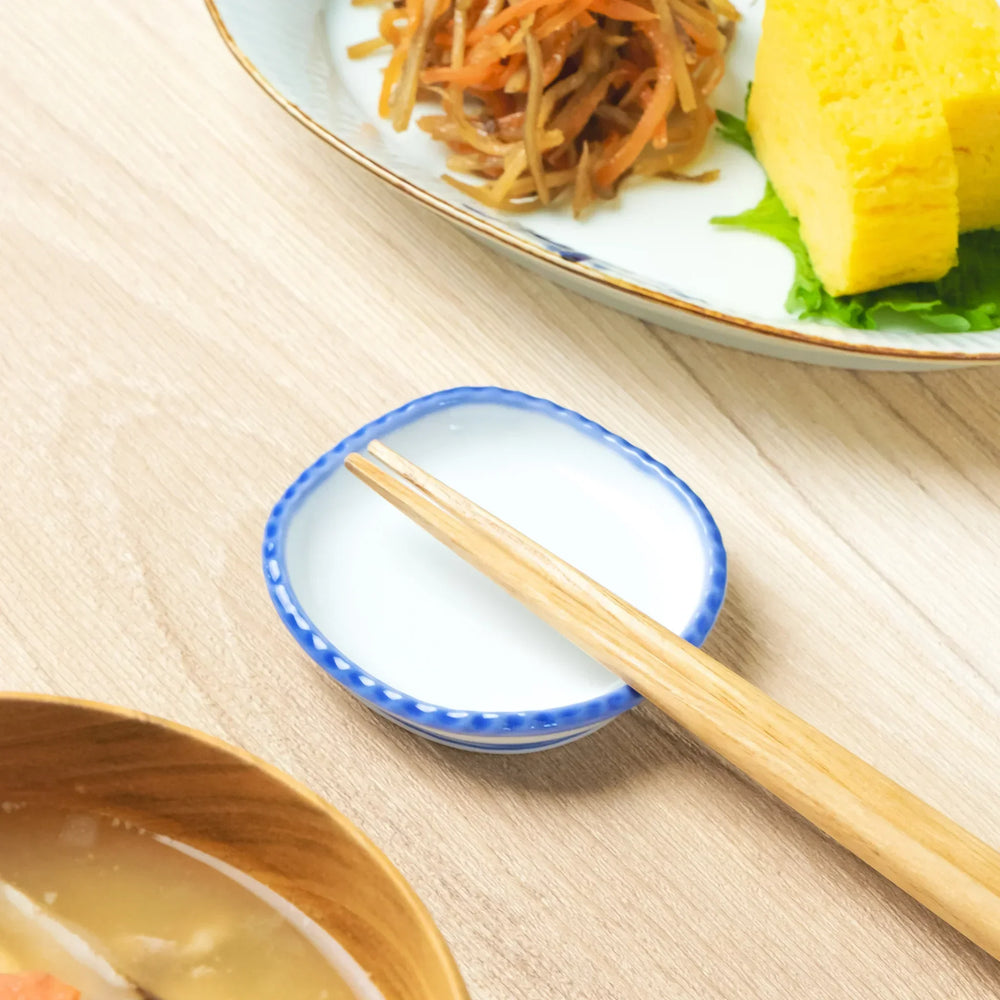 A rounded porcelain Japanese chopstick rest with scalloped edges and a cobalt blue rim, holding wooden chopsticks.