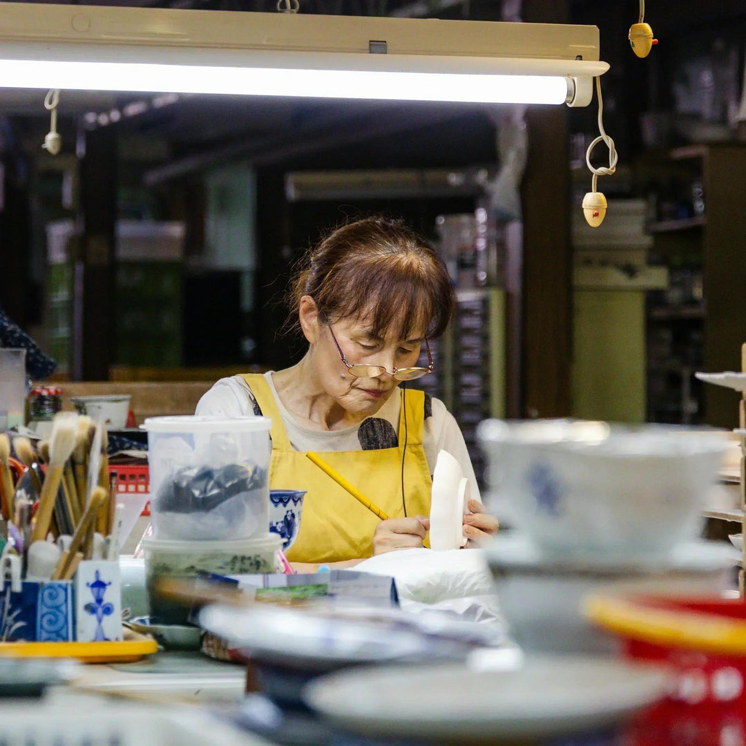 Artisan hand painting porcelain tableware at a Japanese kiln workshop surrounded by brushes and tools.
