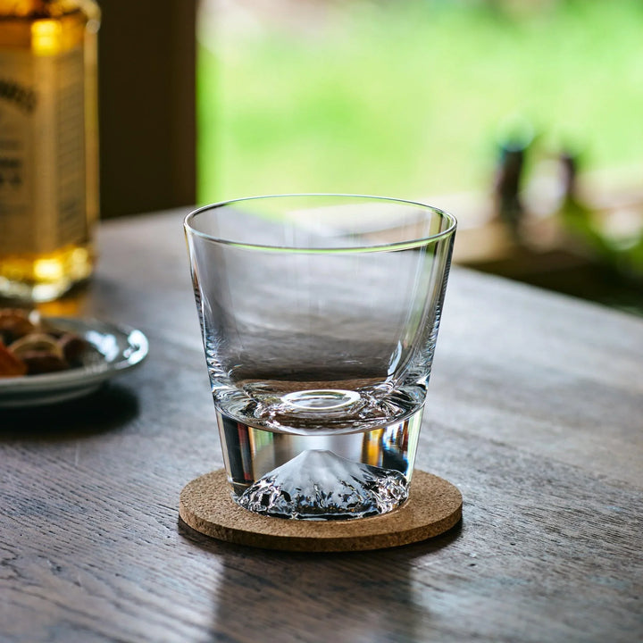 A clear Japanese whiskey glass with a Mt. Fuji-shaped base, placed on a cork coaster on a wooden table.