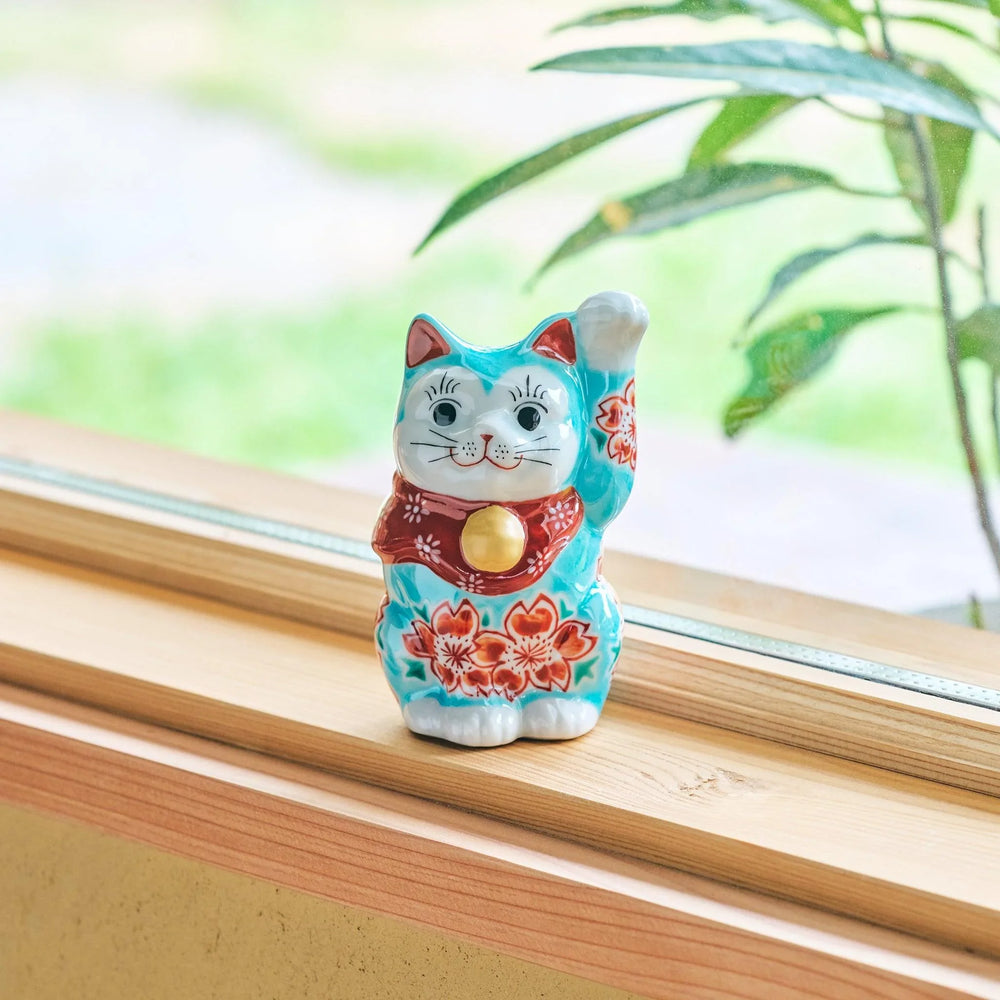 A ceramic maneki-neko (lucky cat) with a turquoise and red floral kimono, raising its paw, displayed on a wooden windowsill.