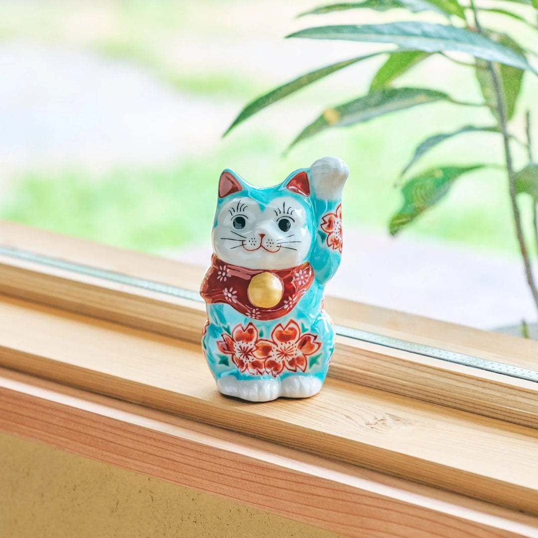 A ceramic maneki-neko (lucky cat) with a turquoise and red floral kimono, raising its paw, displayed on a wooden windowsill.