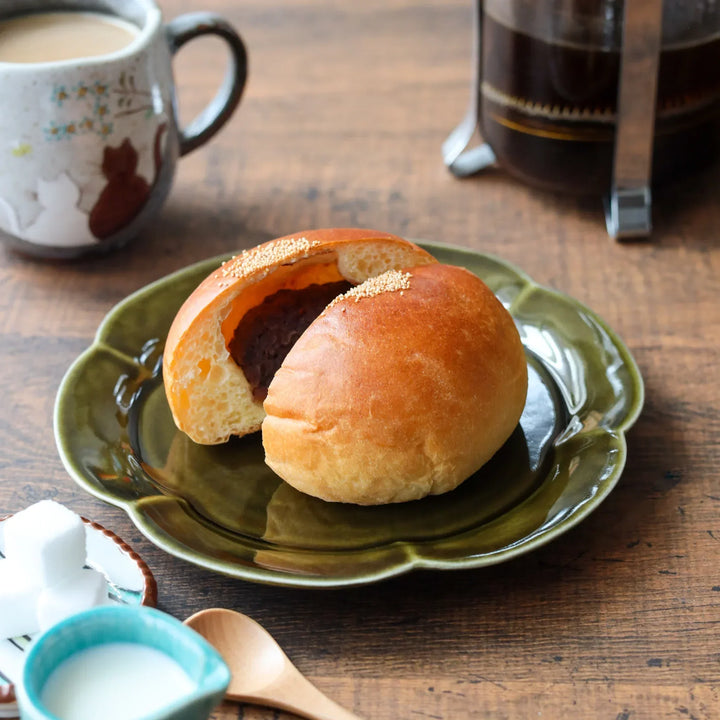 Soft golden-brown anpan bun split revealing red bean paste on a flower-shaped olive-green plate beside a cup of coffee.