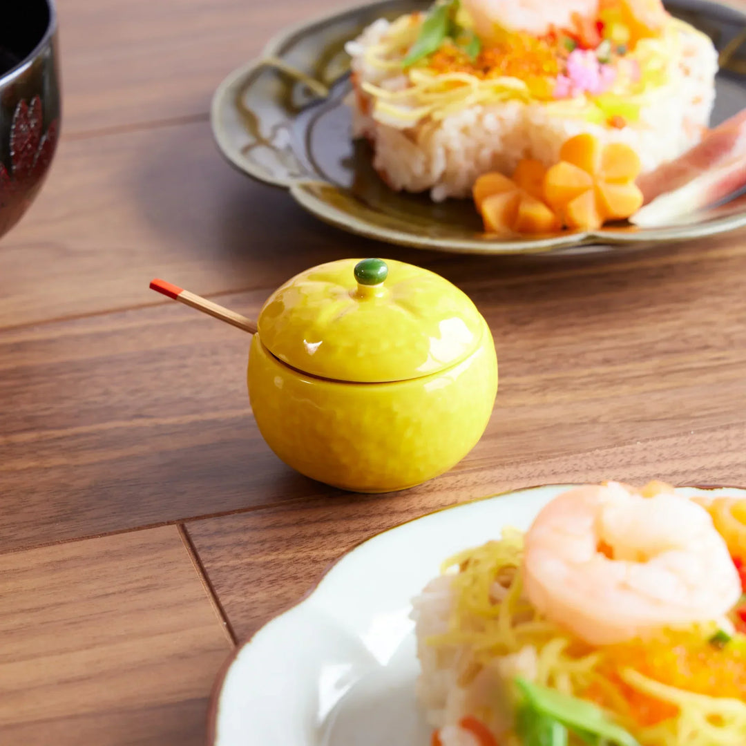 Yellow yuzu-shaped ceramic condiment bowl with lid and tiny spoon, placed beside colorful chirashi sushi.