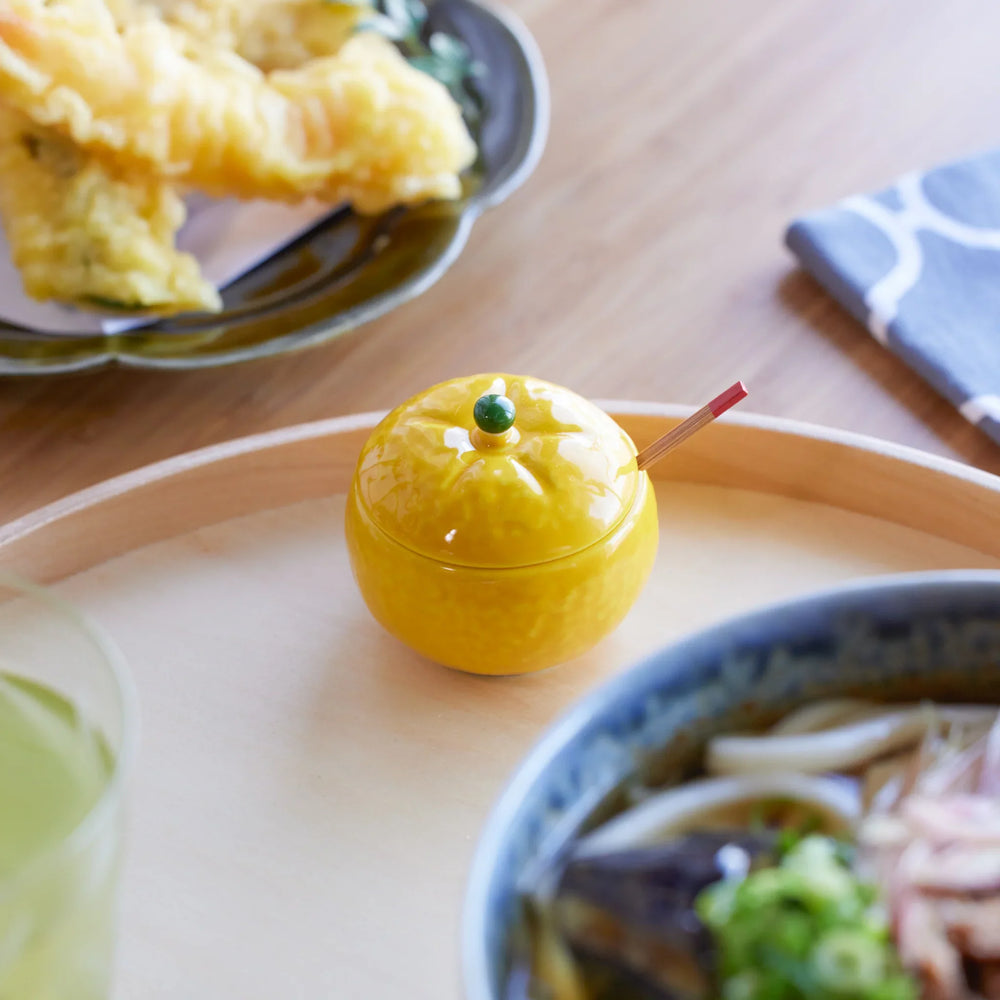 A bright yellow yuzu-shaped condiment bowl with a lid and spoon, placed on a wooden tray next to udon and tempura.