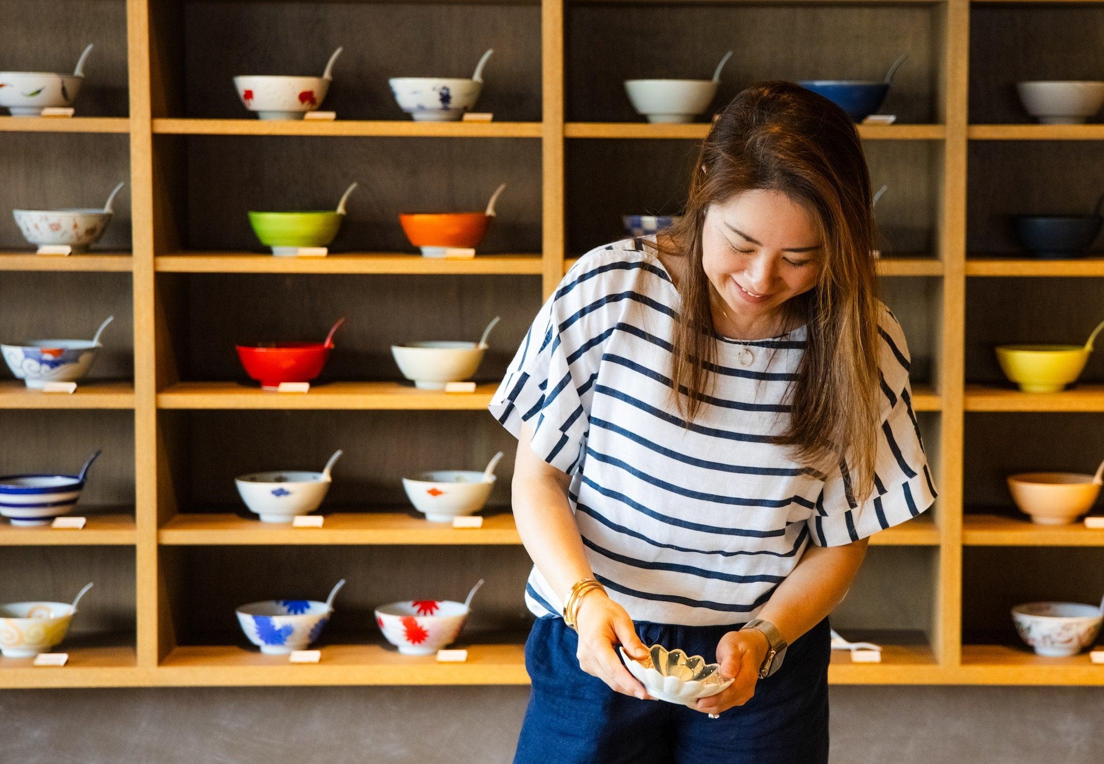 A woman smiling while holding a bowl, standing in front of a shelf filled with colorful bowls of various designs.