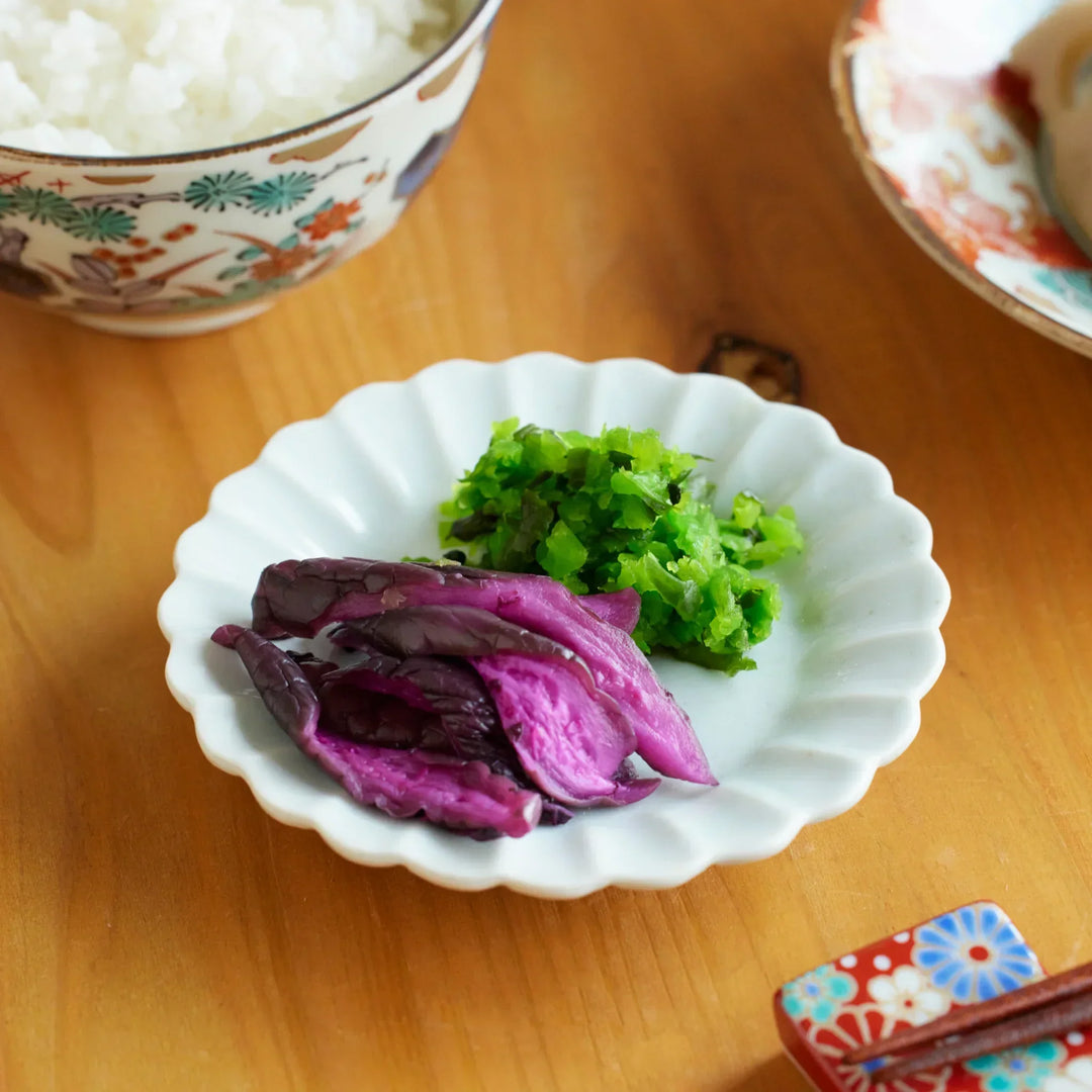 White porcelain chrysanthemum scalloped sauce dish holding purple and green pickles, placed on a wooden dining table.