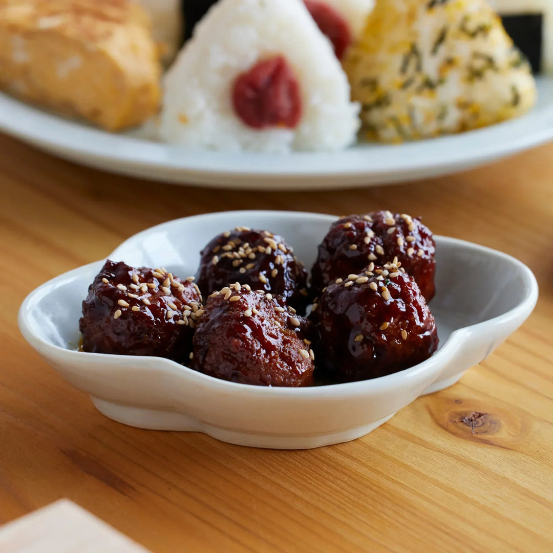 Japanese white porcelain sauce dish with curved rim holding glazed meatballs, shown with onigiri in the background