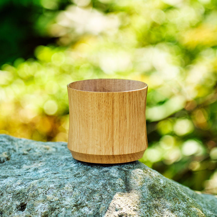 A handcrafted Japanese whiskey cup made from Mizunara oak, resting on a rock in natural outdoor light.