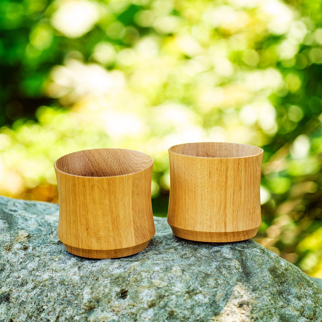 Two Japanese whiskey cups made from Mizunara oak, placed on a rock with soft greenery in the background.