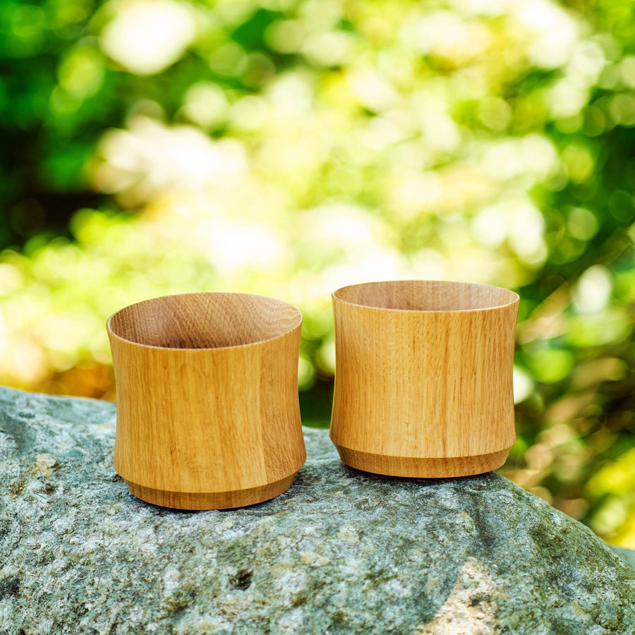 Two Japanese whiskey cups made from Mizunara oak, placed on a rock with soft greenery in the background.