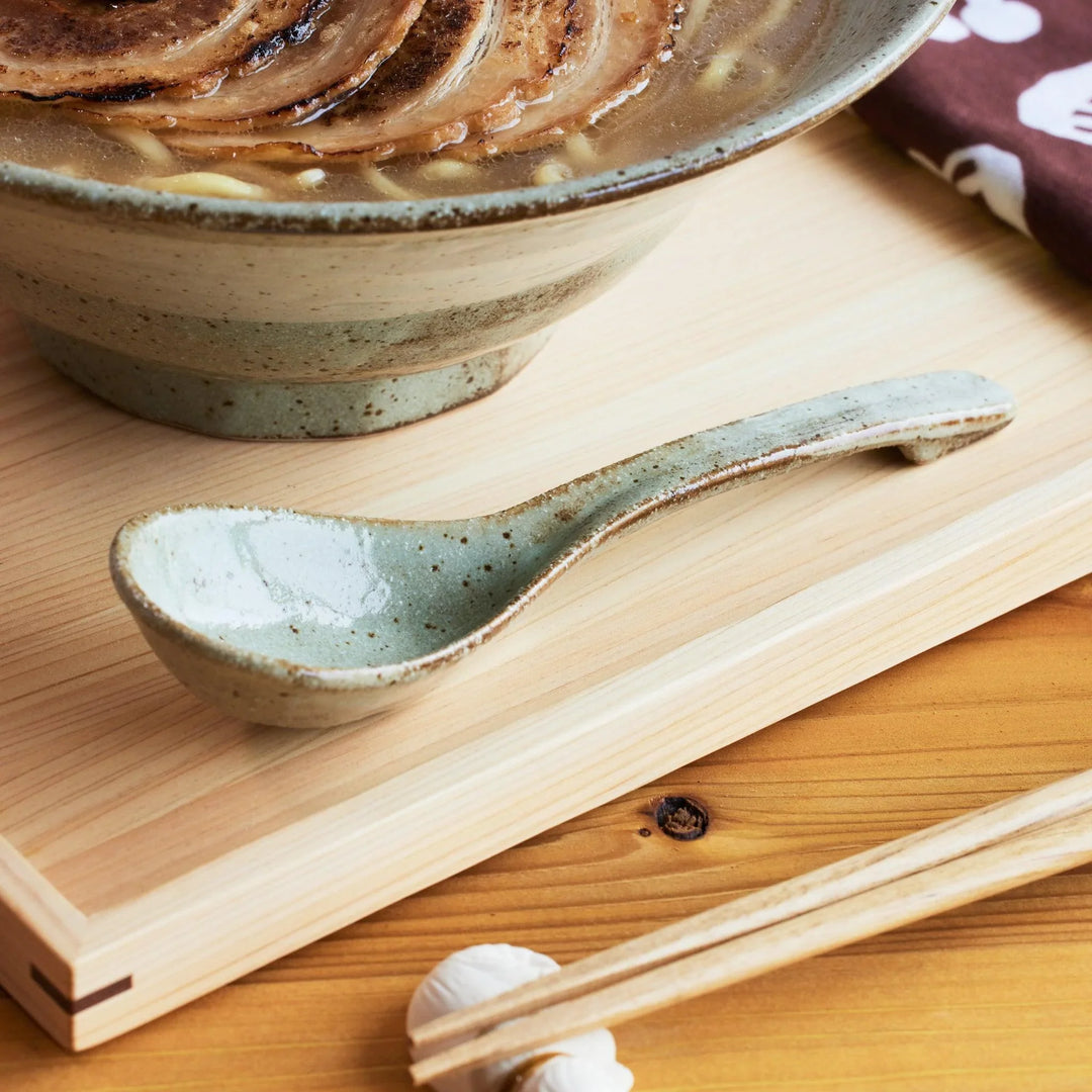 Close-up of a rustic ash glazed ceramic ramen spoon resting on a wooden tray beside a matching ramen bowl.