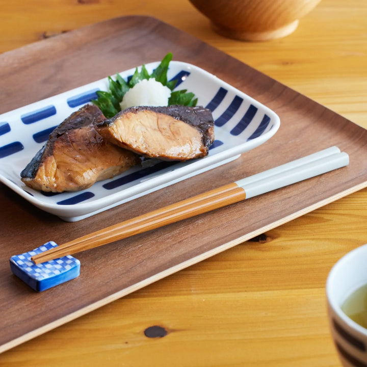 Wood and white chopsticks resting on a blue checkered chopstick rest beside a grilled fish dish on a wooden tray.