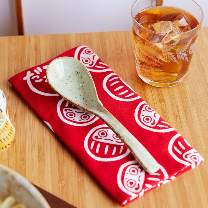 Red tenugui hand towel with white daruma motifs, folded neatly under a ceramic ramen spoon on a wooden table.