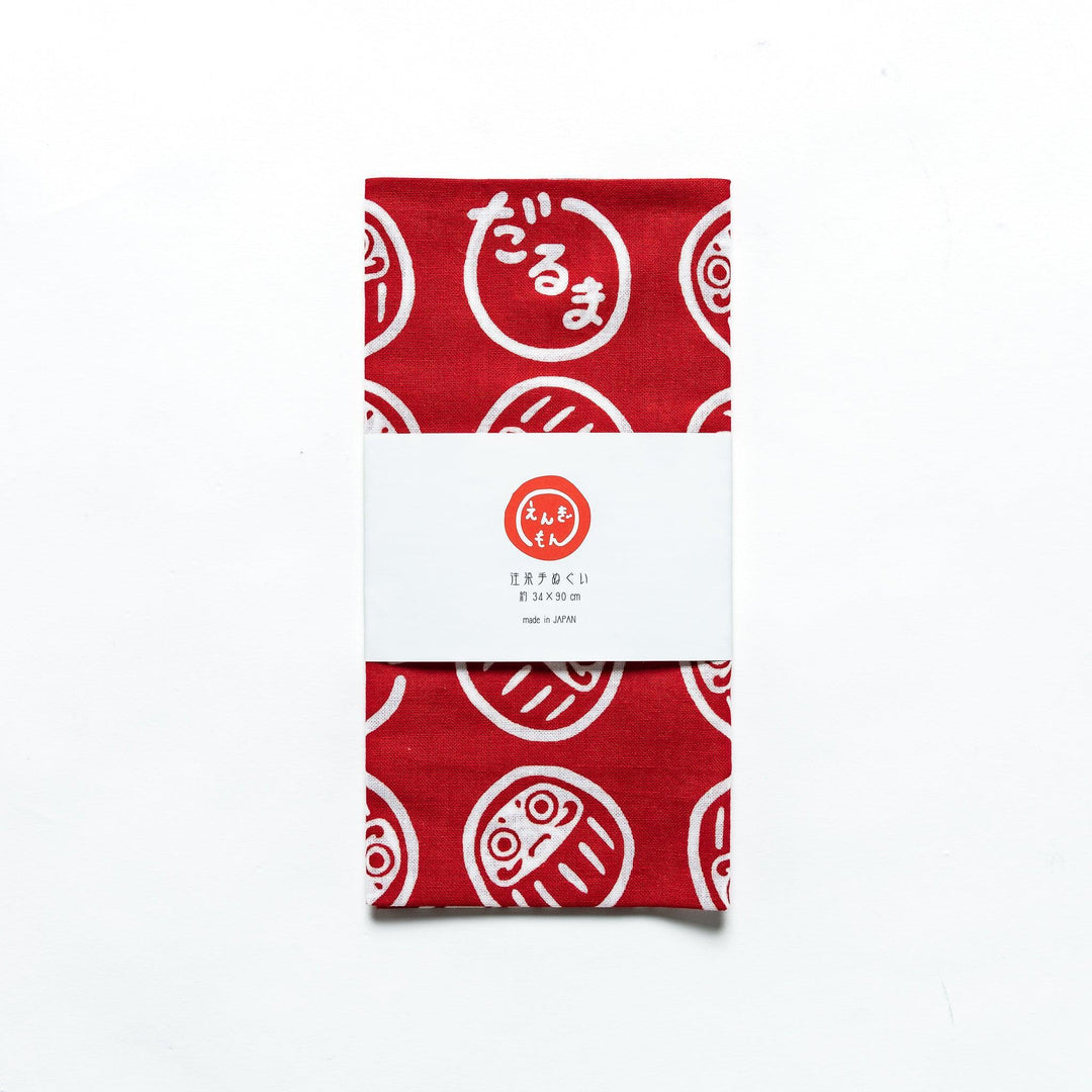 Folded red tenugui with white Daruma patterns, enclosed in a product sleeve, placed on a white background.